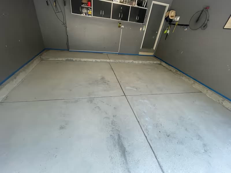 Bare concrete slab masked with blue painter's tape along every baseboard before coating